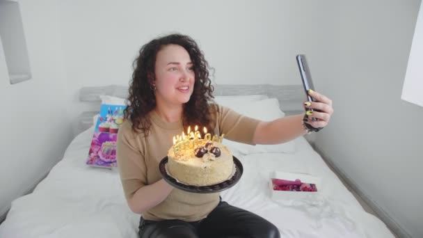 Fiatal lány egy ágyban egy szállodában, kommunikál és ünnepli a születésnapját online és gyertyákat fúj el egy tortán — Stock videók