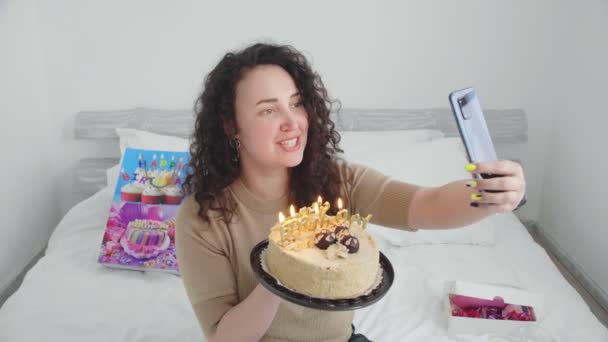 Fiatal lány egy ágyban egy szállodában, kommunikál és ünnepli a születésnapját online és gyertyákat fúj el egy tortán — Stock videók