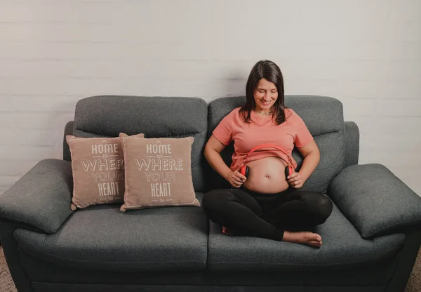 Bebek müzik dinleyebilsin diye kanepeye oturmuş karnına kulaklık takan hamile bir kadın. Bebek için yeni duygular kavramı. Boşluğu kopyala