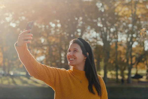 Portrait of a young Latina woman smiling while taking a selfie or making a video call, in a park in autumn. Copy space.