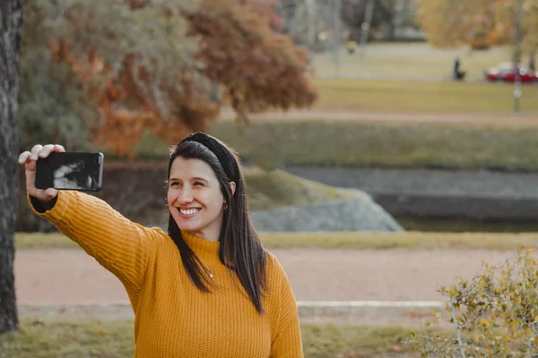 Portrait of a young Latina woman smiling while taking a selfie or making a video call, in a park in autumn. yellow and green contrast. Copy space.