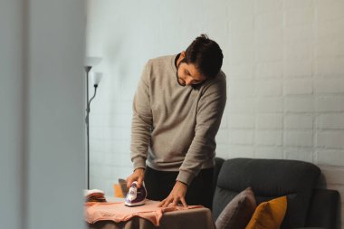 Latin bir adam cep telefonuyla konuşurken kadın kıyafetlerini ütülüyor. Genç yetişkin ev işleri yapıyor. Cinsiyet eşitliği kavramı.