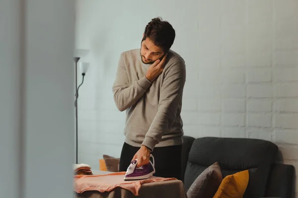 Latin bir adam cep telefonuyla konuşurken kadın kıyafetlerini ütülüyor. Genç yetişkin ev işleri yapıyor. Cinsiyet eşitliği kavramı.