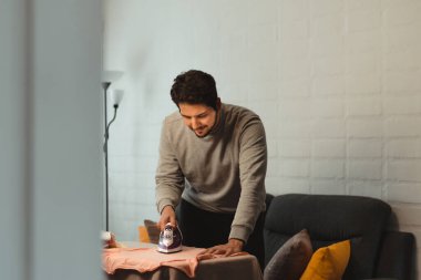 Latin adam kadın kıyafetlerini oturma odasında ütülüyor. Giysileri yakından gözlemler ve gülümser ve anın tadını çıkarır. Evde eşit ev işleri kavramı.