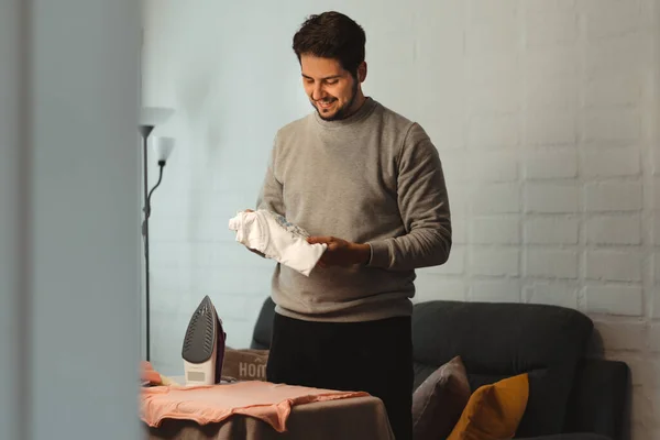 Latin man with braces ironing women's clothes in living room. Standing holding an outfit and smiling. Concept of equal chores at home.