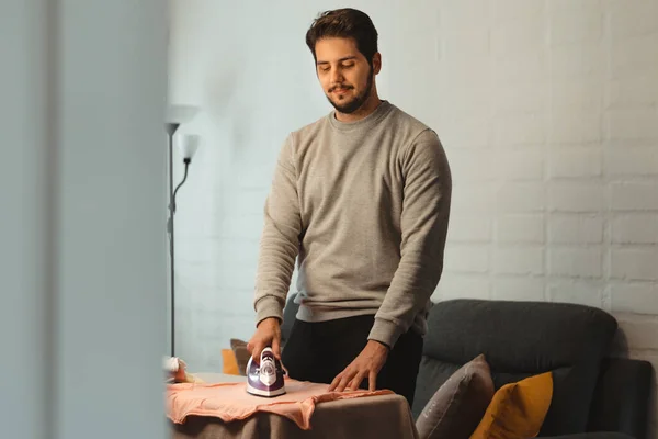 Latin man ironing women's clothes in the living room. Standing looking down smiling. Concept of equal chores at home.