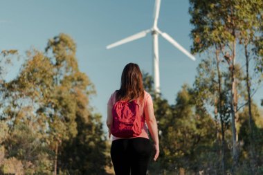 Kırmızı sırt çantalı İspanyol bir kadının yatay görüntüsü. Doğanın ortasında yürüyor. Yel değirmenine doğru ilerliyor. İyi sağlık ve eğlence sporu kavramı.