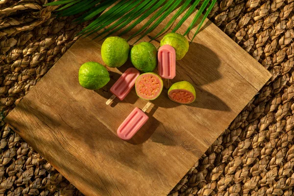 Tahta tabanlı meyveli Guava dondurması, güneş ışığı, palmiye yaprağı, üst manzara.