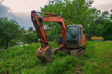 Front view: modern caterpillar excavator with bucket lowered stands on green grass. In the background are trees and the evening sky.