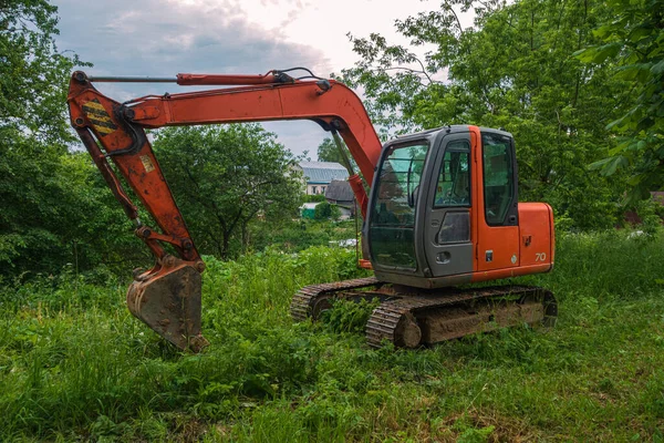 A modern excavator stands on the grass. The bucket is lowered. In the background, trees and a village house, the evening sky.