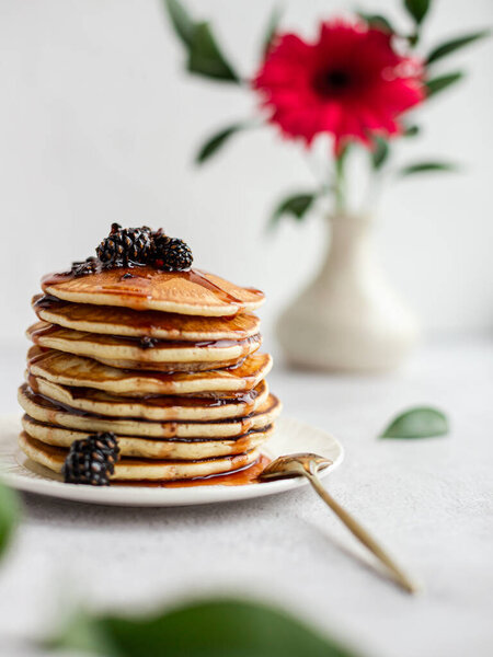 A stack of pancakes with jam from cones on a light background. In the background there is a flower in a vase