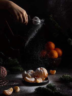Yeni yıl kompozisyonu ve tabakta mandalina. Çam kozalakları. Siyah arka plan. Şenlik dekorasyonu. Bir el mandalinalara pudra şekeri döker.