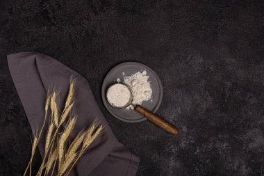 Siyah arka planda beton bir tabakta un bulunan ahşap saplı bir süzgeç. Buğday kulakları ve keten peçete. Pişirme hazırlığı