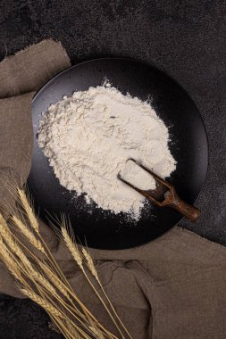 Un kepçesiyle bir tabakta un. Siyah arkaplanın dokusu. Buğday kulakları. Gri keten peçete