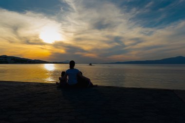 Mother and her daughters watching the sunset and the silhouette of a fishing boat.A family loving moment against a dramatic sky.