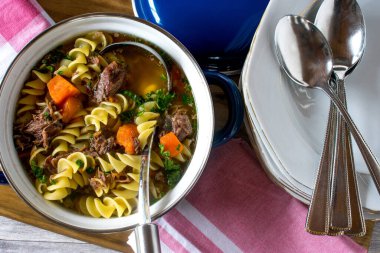 Mutfak arka planında tabak ve kaşıklarla servis edilen mavi bir tencerede taze pişmiş etli şehriye çorbası.