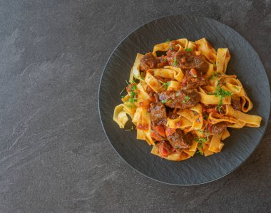 Makarna ve soslu lezzetli sığır eti yahnisinin genel manzarası. Domateslerden, otlardan ve sebzelerden yapılmış ve siyah bir tabakta fotokopi alanı ile servis edilmiş.