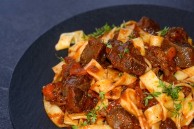 Biftek bacakları, kök sebzeleri, domatesler ve koyu bir tabakta makarnayla servis edilen taze pişmiş sığır eti yahnisinin yakın görüntüsü.