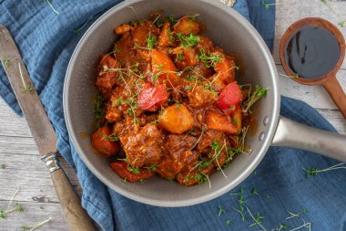 Patates ve sebzeyle pişirilmiş baharatlı domuz yahnisi ahşap masa arkasında balık soslu bir tavada servis ediliyor.