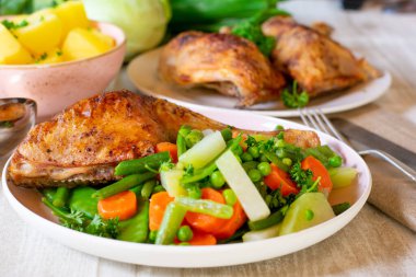 Roasted chicken legs with mixed vegetables such as carrots, green peas, green beans, kohlrabi, sugarsnap peas served on a plate with table background. 