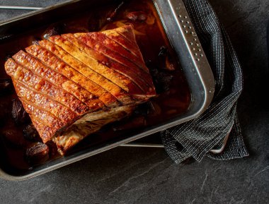 Roast pork belly with delicious golden crust  in a baking pan with red onions isolated on dark table background with copy space. Overhead and closeup view