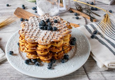 Beyaz tabakta yabanmersini ve pudra şekerle servis edilen taze pişmiş waffle yığını. Sağlıklı organik khorasan ya da kamut unundan yapılmış gözlemeler yukarıdan fotokopi alanı ile birlikte rustik ahşap masa zemininde servis edilir. 