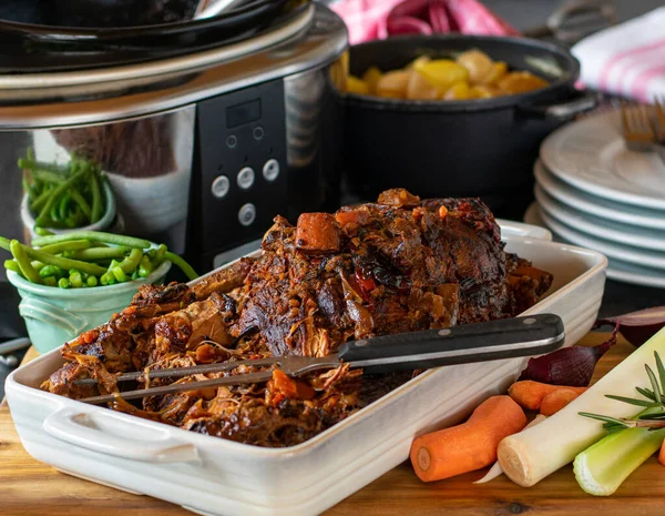 Slow cooker roast pork with gravy and vegetables served with a meat fork in a baking dish with kitchen background