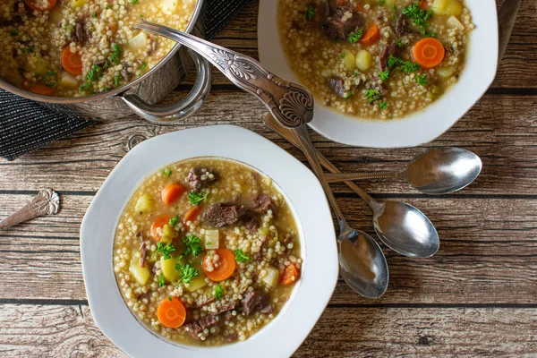 fresh and homemade cooked barley soup with beef and root vegetables served on rustic and white plates with pot on a dinner table. Overhead view