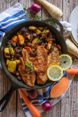 Kızarmış kırmızı balık ve patates, havuç, soğan ve havuç gibi kızarmış sebzelerle taze ve ev yapımı balık yemeği yukarıdan gelen sade ve ahşap bir masa arka planında otlar ve limonla servis edilir.