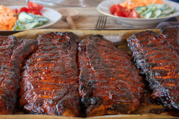Fresh and homemade cooked marinated and honey glazed spareribs served on a baking tray with plates of salad in the background. Closeup view