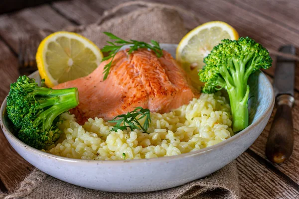 Plate with risotto , grilled fish and vegetables. Fresh cooked fish dish with grilled salmon fillet served with creamy risotto and fresh steamed broccoli on a rustic and wooden table. Closeup and isolated view with blurred background