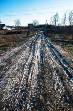Kışın kırsal alanda karlı çamurlu kırsal yol. Yüksek kaliteli fotoğraf, buzlu çamurlu köy yolu. Sakin bir köy boyunca giden lastik izleri. Kış yolculuğunu, altyapıyı ve günlük kırsal yaşamı yansıtıyor..