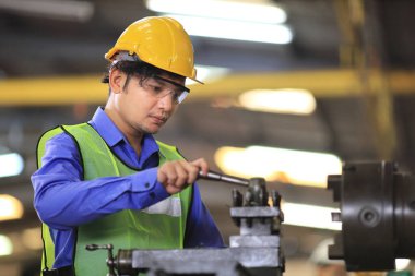 Güvenlik kaskı ve yansıtıcı bez giyen Asyalı tamirci fabrikanın içinde torna makinesiyle çalışıyor.