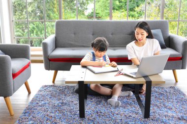 Asyalı anne evde bilgisayarla çalışırken kızının evde eğitim ve eğitim konsepti için okuldan sanat sınıfı ödevi yapmasını izliyor.