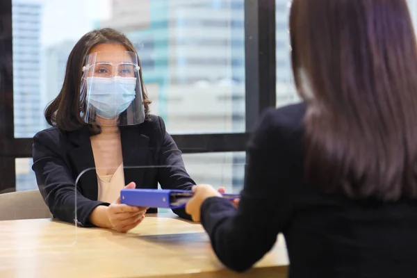 Manager from HR department wearing facial mask is interviewing new applicant who is handing her resume and profile through the partition for social distancing, transaction and new normal policy