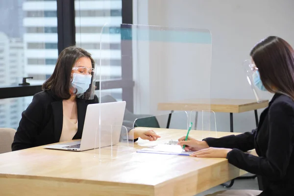 Manager from HR department wearing facial mask is interviewing new applicant who is signing her contract through the partition for social distancing and new normal policy