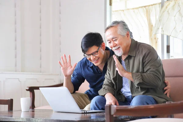 Elder Asian man and his son using video call from computer to say hi to the rest of his family in his cozy retirement home