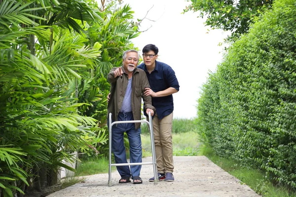 Happy elder Asian man using walker while walking for exercise around the garden with his son taking care of him at the retirement age with copy space