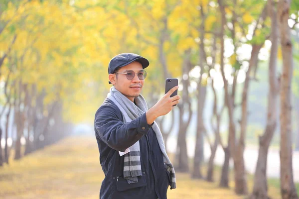 Süveter giyen Asyalı adam, bahar mevsiminde sarı çiçek ağacıyla parkta selfie çekmek için cep telefonu kullanıyor.