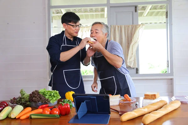 Old Asian man in retirement age spending time to learning how to cook healthy food from internet with his son via digital tablet using variety of organics vegetables with copy space