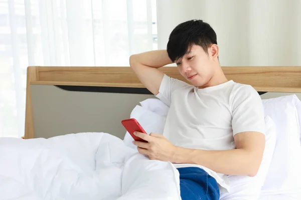 Asian man wearing white cloth using mobile phone for checking morning news and social media right after he woke up on his bed with copy space