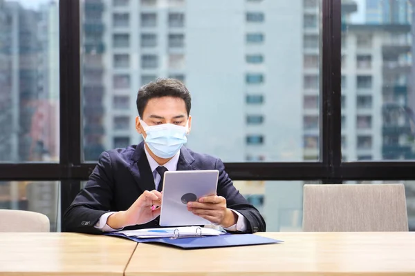 Asian male employee in formal suit wearing protective facial mask working online with tablet in business office during coronavirus or covid-19 pandemic as new normal and social distancing policy