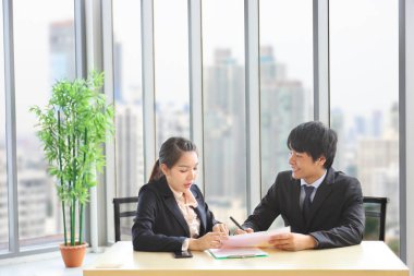 Resmi takım elbise giyen Asyalı işadamları, resmi iş ofisindeki yıllık rapor üzerinde tartışıyor. Arka planda modern şehir manzarası ve fotokopi alanı var.