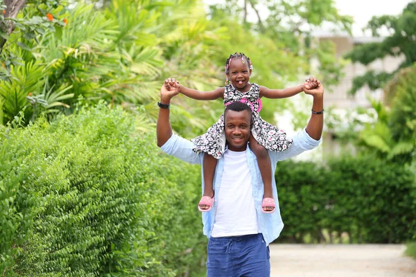 アフリカ系アメリカ人の父親は 近所の家の外を歩いている間に 幸せな精神的健康と楽しい時間を一緒に過ごすことで 彼の小さな娘にピギーバックに乗ることを与えました — ストック写真