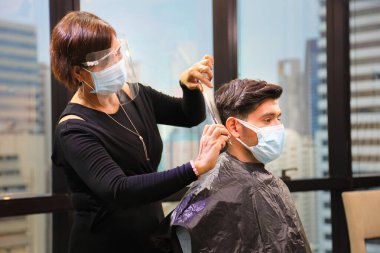 Kuaförde yüz maskesi takan berber ve müşteri, Coronavirus veya covid-19 salgınından sonra yeni normal ve sosyal mesafeli yaşam tarzlarında salgından kaçınmak için saç tıraşı olurken,