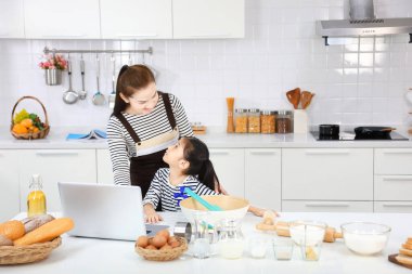 Mutlu Asyalı anne, genç kızına bilgisayardan tarifine bakarak beyaz mutfakta ekmek pişirmeyi öğretiyor.