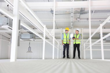 İki Asyalı müteahhit güvenlik ekipmanı giyip deponun içindeki fabrikanın gelecekteki genişleyen iş planlarını tartışıyor ve denetliyor.