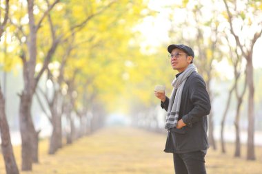 Asyalı bir adam elinde kahve fincanıyla sonbahar mevsiminde halka açık parkta yürürken yaprak fotokopi alanı ile sarıya dönüşüyor.