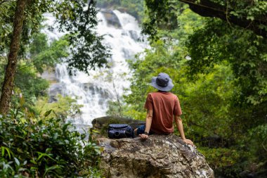 İnsanlar rahatça oturup tropikal ormanlık alanlarda meditasyon ve farkındalık için şelaleler eşliğinde huzur dolu alternatif terapi ormanı ve dinlendirici doğa sesini dinliyorlar.