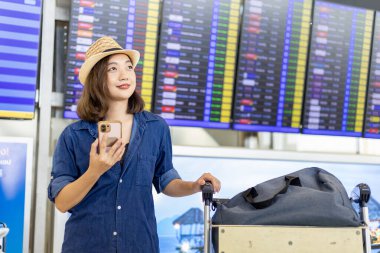 Bagajı olan genç Asyalı kadın yolcu, uluslararası seyahat ve tatil uçuşu için havaalanı terminalindeki bilet kontrolünden sonra uçuş kartına bakıyor.